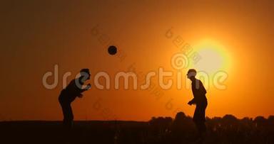 两个男孩在日落时踢足球。 日落时孩子们玩球的剪影。 幸福家庭的概念
